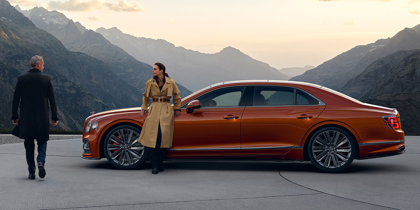 Bentley Zug Bentley Flying Spur Speed parked in Orange Flame coloured exterior parked, with mountainous background and two people in view.