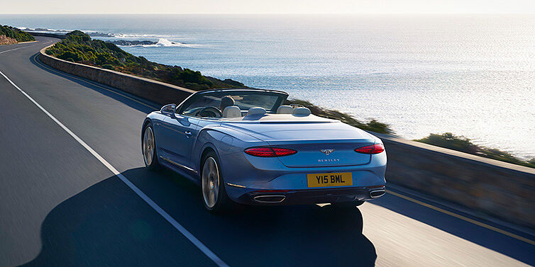 Bentley Zug New Bentley Continental GTC Azure convertible rear view in Light Windsor blue paint with 22 inch ten spoke wheels driving dynamically on an ocean road in the sunshine