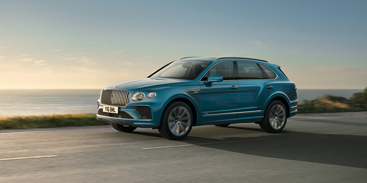 Bentley Zug Bentley Bentayga Azure SUV in Topaz blue paint driving dynamically by the ocean with 22 inch 10 spoke directional wheels