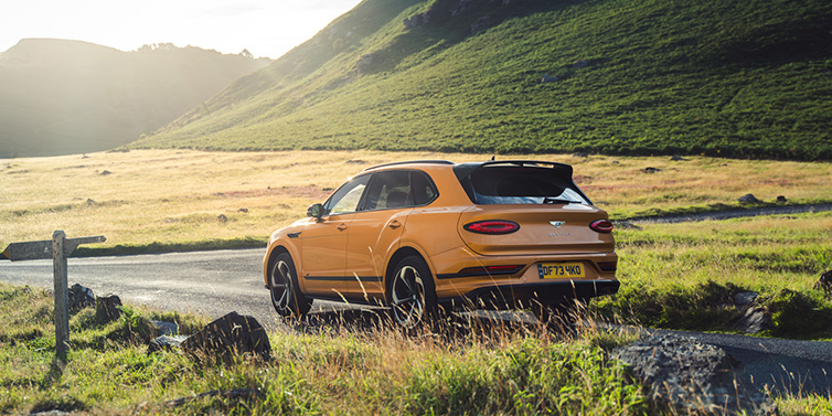 Bentley Zug Bentley Bentayga S SUV rear three quarter in Sunburst Gold paint on a road surrounded by green hills