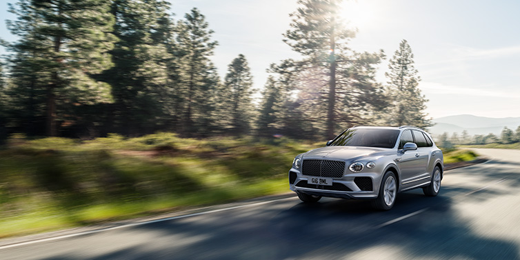 Bentley Zug Bentley Bentayga SUV front three quarter in Moonbeam silver paint driving dynamically along a tree-lined road