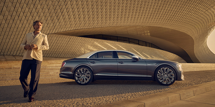 Bentley Zug Bentley Flying Spur Mulliner sedan in Rose Gold over Magnetic duotone paint parked by a curved wall with person in suit jacket standing on the left